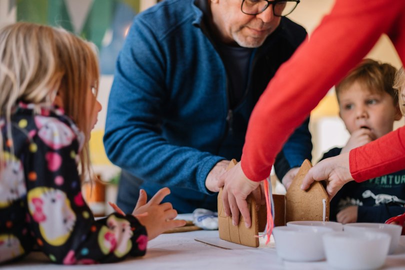 Photo with two people making a gingerbread house and three children looking on.