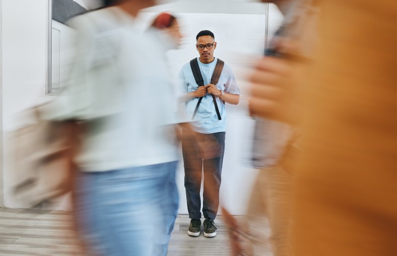 Young man in the background, looking sad as blurred figures walk past, ignoring him.